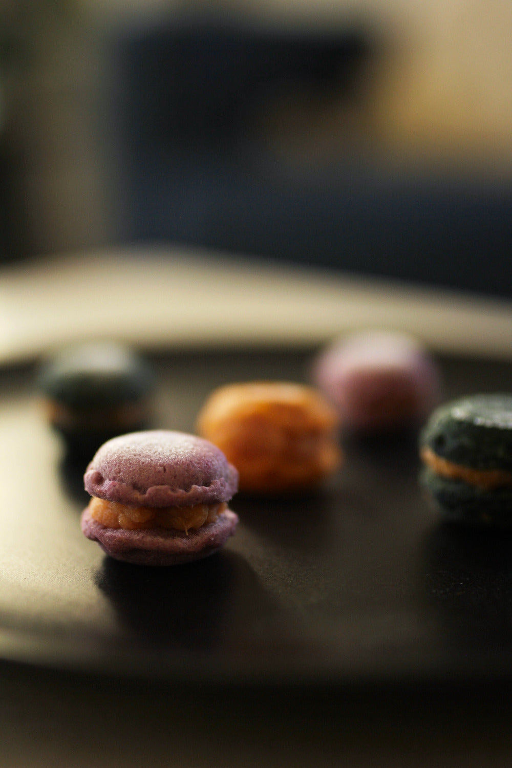 Four colorful French macarons with orange cream filling arranged on a dark plate, photographed in a shallow depth of field with a blurred background.