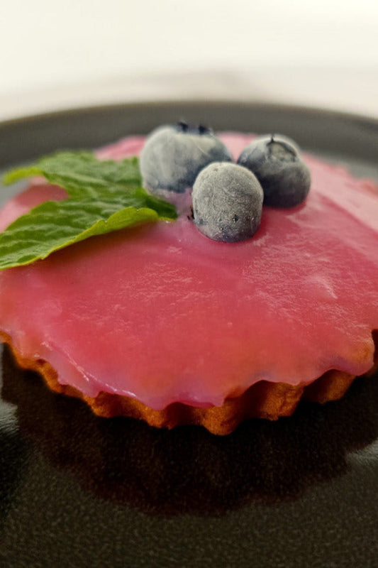 A small pink berry tart or tartelette with a fluted pastry crust, topped with three blueberries and a mint leaf, served on a glossy black plate.