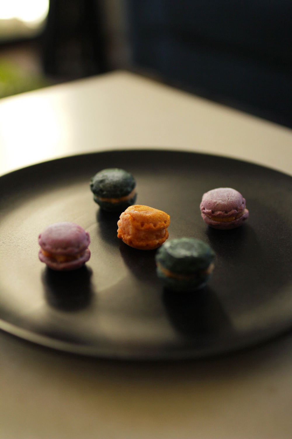 Four colorful French macarons in purple, orange, and dark green, arranged on a matte black plate.
